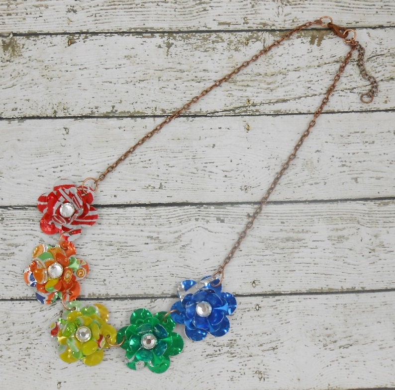 May include: A handmade necklace featuring six colorful flower pendants made from recycled materials. Each flower is a different color, including red, orange, yellow, green, and blue, and has a sparkling gem in the center. The chain is a copper color.