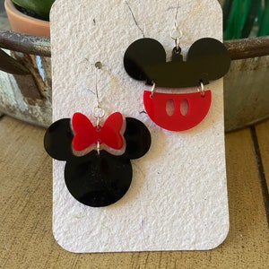 May include: A pair of acrylic earrings in black and red, depicting Mickey Mouse and Minnie Mouse. One earring shows Mickey's head with red shorts, the other Minnie's head with a red bow. They hang from silver-coloured hooks.