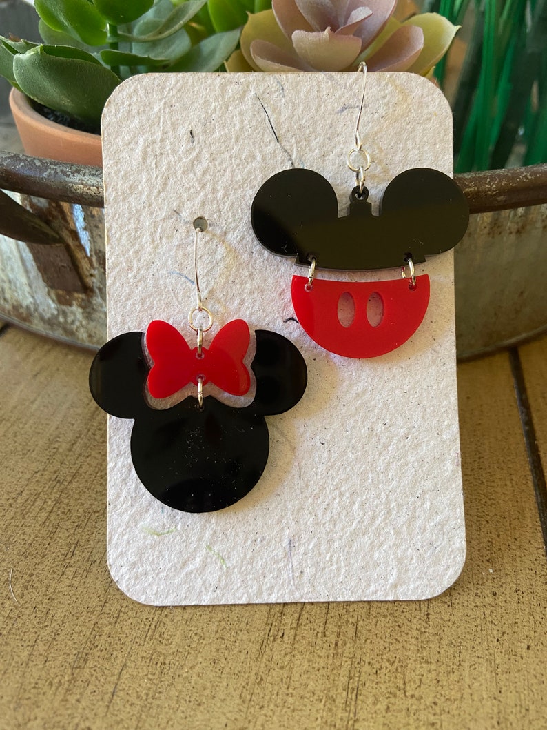 May include: A pair of acrylic earrings in black and red, depicting Mickey Mouse and Minnie Mouse. One earring shows Mickey's head with red shorts, the other Minnie's head with a red bow. They hang from silver-coloured hooks.