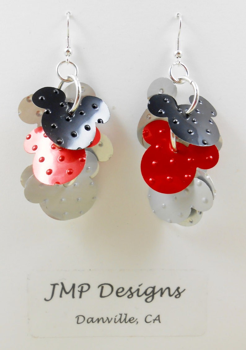 Puede incluir: Pendientes colgantes con un dise&ntilde;o en capas de Mickey Mouse. Los pendientes presentan elementos met&aacute;licos rojos, negros y plateados con un acabado texturizado y punteado. Cuelgan de ganchos plateados.