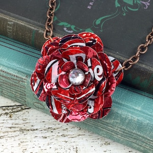 May include: A handmade necklace featuring a large, red flower pendant crafted from recycled aluminum cans. The flower has a sparkling rhinestone center and is attached to a copper-colored chain. The pendant is approximately 3 inches in diameter.