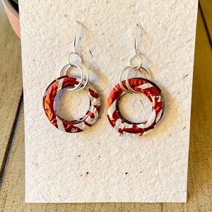 May include: A pair of handmade earrings featuring circular red and white patterned pendants. The pendants are connected to silver-colored hoops and ear wires. The earrings are displayed on a textured, off-white surface.