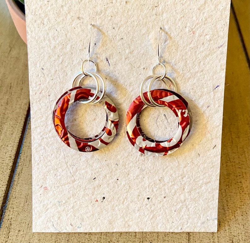 May include: A pair of handmade earrings featuring circular red and white patterned pendants. The pendants are connected to silver-colored hoops and ear wires. The earrings are displayed on a textured, off-white surface.