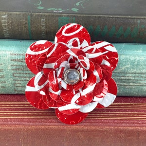 May include: A handmade flower brooch crafted from recycled red and white aluminum. The petals are cut from a soda can, with a clear gem in the center. The brooch is a unique accessory.
