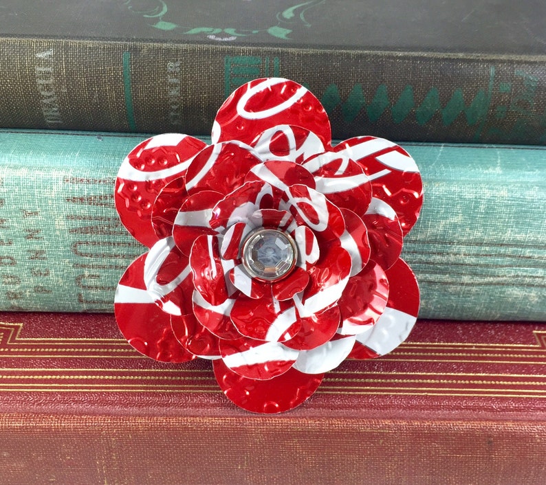 May include: A handmade flower brooch crafted from recycled red and white aluminum. The petals are cut from a soda can, with a clear gem in the center. The brooch is a unique accessory.