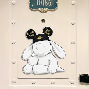 May include: A white door decorated with nautical-themed cutouts, including anchors, life preservers, and Mickey Mouse silhouettes. A cartoon bunny wearing a hat is affixed to the door. The door has a room number plaque that reads "10186".