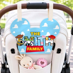 May include: A light blue stroller sign shaped like mouse ears with white clouds, featuring the text THE SMITH FAMILY in a Toy Story-inspired font. The sign hangs from the stroller handle, while a plush rabbit and a pink toy sit in the pocket.