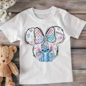 Pu&ograve; includere: T-shirt bianca con un cartone animato di un personaggio alieno blu, un castello e un fiocco. Il fiocco &egrave; rosa e blu a pois. La parola "NAME" &egrave; scritta sul disegno.