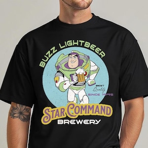 May include: Black t-shirt featuring a cartoon of Buzz Lightyear holding a beer mug. The design includes the text "BUZZ LIGHTBEER" above the character and "STAR COMMAND BREWERY" below. The design is set on a light blue circle.