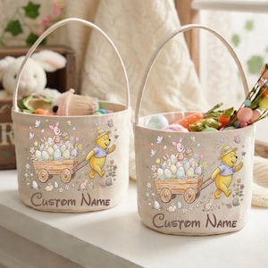 May include: Two beige Easter baskets with white handles. Each basket features a Winnie the Pooh design with a wagon of Easter eggs and the text "Custom Name". One basket is filled with carrots and decorative flowers.