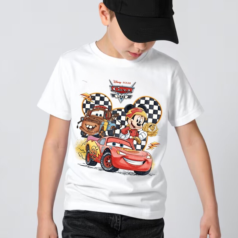 May include: A white short-sleeve t-shirt featuring a graphic print of Disney Pixar Cars Land. The design includes Lightning McQueen, Mater, and Mickey Mouse wearing a racing outfit, set against a checkered background with the Cars Land logo.