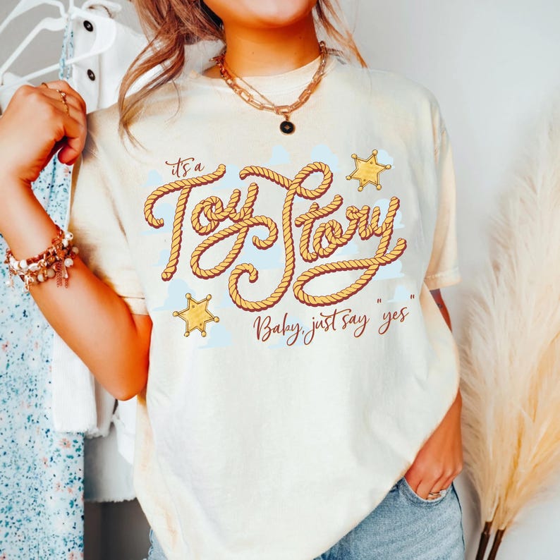 Peut inclure: Un t-shirt &agrave; manches courtes de couleur cr&egrave;me avec un motif graphique portant le texte "it's a Toy Story" &eacute;crit dans une police style corde, accompagn&eacute; de deux ic&ocirc;nes d'&eacute;toiles de sh&eacute;rif jaunes et du texte "Baby, just say yes".