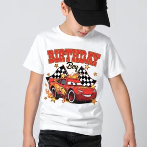 Puede incluir: Una camiseta blanca de manga corta con un gr&aacute;fico de un coche de carreras rojo con el n&uacute;mero 95, banderas a cuadros, llamas naranjas y estrellas amarillas. El texto "BIRTHDAY Boy" est&aacute; impreso en letras rojas a cuadros sobre la ilustraci&oacute;n.