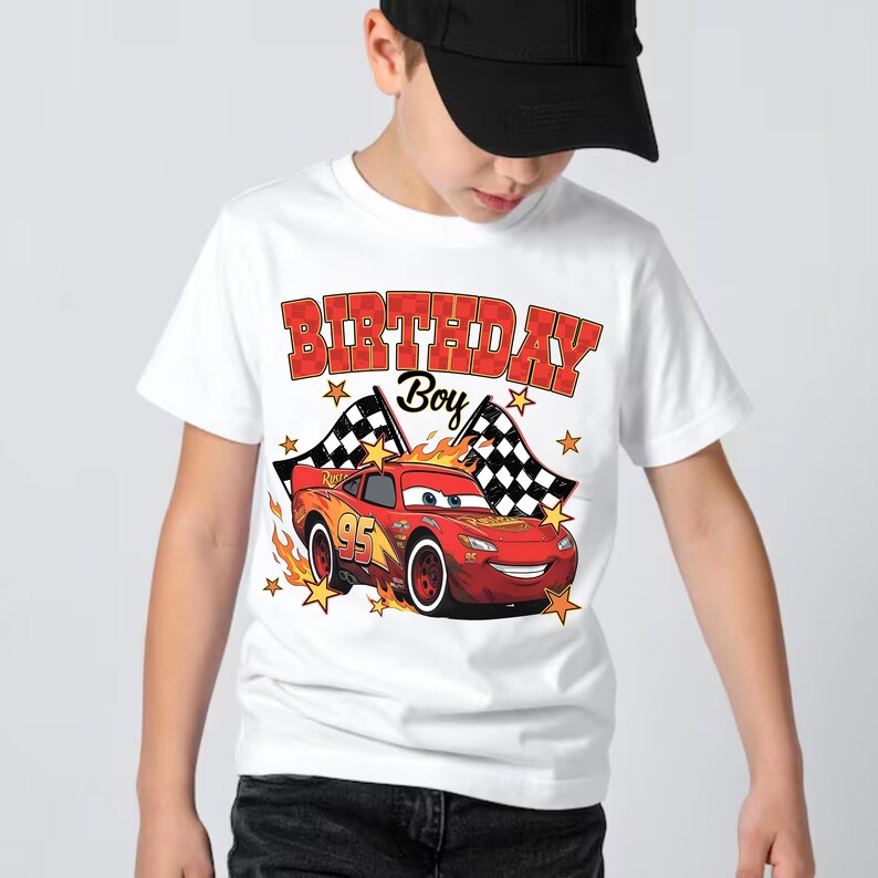 Puede incluir: Una camiseta blanca de manga corta con un gr&aacute;fico de un coche de carreras rojo con el n&uacute;mero 95, banderas a cuadros, llamas naranjas y estrellas amarillas. El texto "BIRTHDAY Boy" est&aacute; impreso en letras rojas a cuadros sobre la ilustraci&oacute;n.
