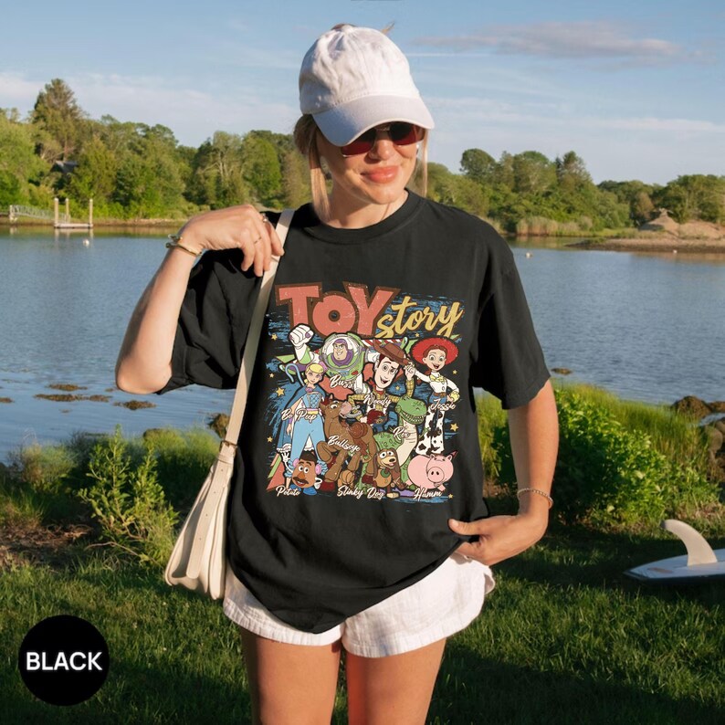 Puede incluir: Camiseta negra con un gr&aacute;fico colorido de los personajes de Toy Story y el texto "Toy Story". La camiseta la lleva una persona en un entorno natural al aire libre. La persona lleva una gorra blanca y gafas de sol.