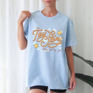 Peut inclure: Un t-shirt bleu clair &agrave; manches courtes avec une impression graphique du texte "It's a Toy Story" &eacute;crit dans une police style corde, accompagn&eacute; de deux ic&ocirc;nes d'&eacute;toiles de sh&eacute;rif jaunes et du texte "Baby, just say yes" en marron.