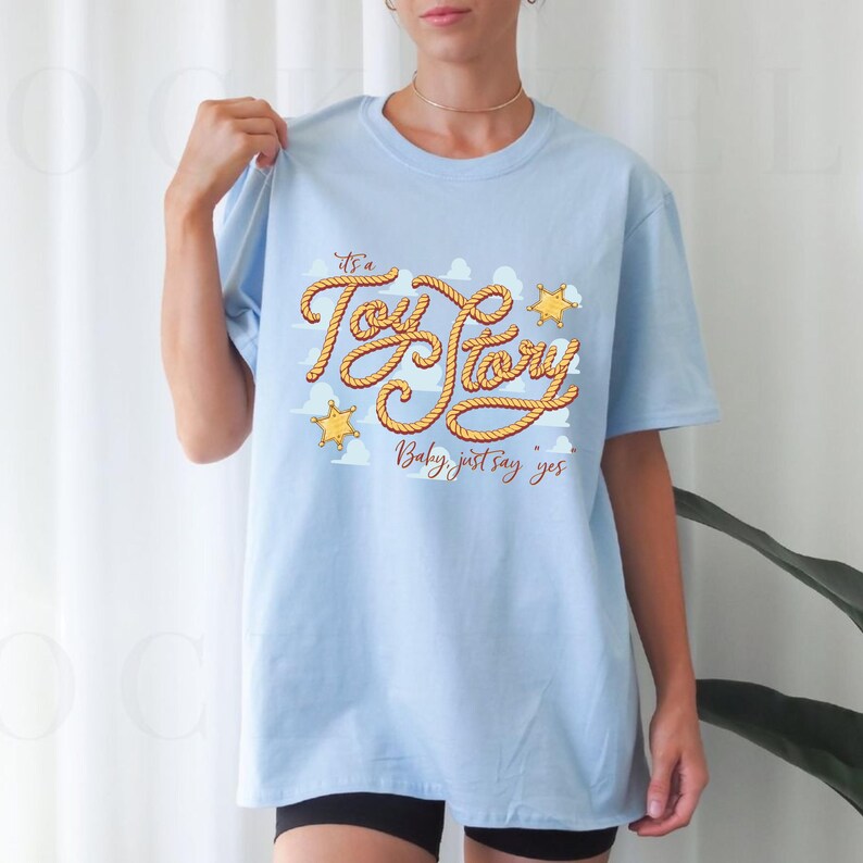 Peut inclure: Un t-shirt bleu clair &agrave; manches courtes avec une impression graphique du texte "It's a Toy Story" &eacute;crit dans une police style corde, accompagn&eacute; de deux ic&ocirc;nes d'&eacute;toiles de sh&eacute;rif jaunes et du texte "Baby, just say yes" en marron.