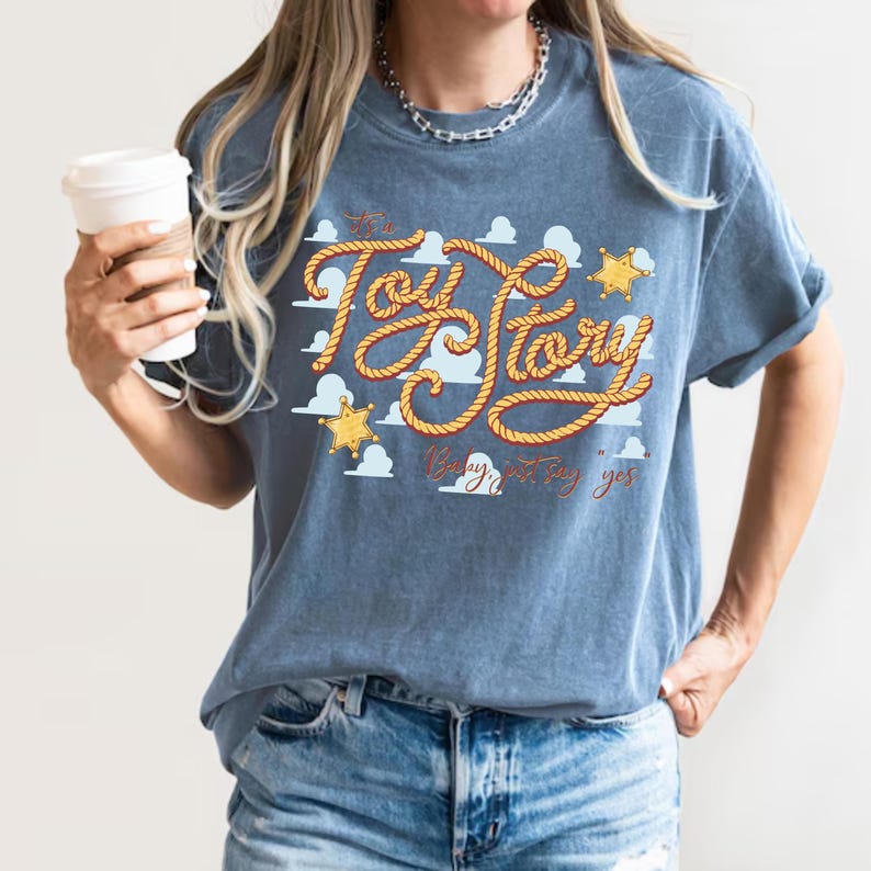 Peut inclure: Un t-shirt bleu &agrave; manches courtes avec une impression graphique du texte "It's a Toy Story" &eacute;crit dans une police style corde, accompagn&eacute; d'ic&ocirc;nes d'&eacute;toiles de sh&eacute;rif jaunes et de nuages blancs, avec le texte additionnel "Baby, just say yes".