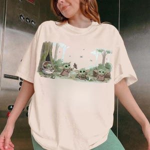 Puede incluir: Camiseta color crema con una ilustraci&oacute;n de dibujos animados de Grogu de The Mandalorian. El dise&ntilde;o muestra a Grogu en varias poses con un fondo de bosque. La palabra "cool" est&aacute; escrita en la camiseta.