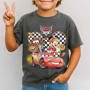 May include: A charcoal gray short-sleeve t-shirt featuring a graphic print of Disney Pixar Cars Land. The design includes Lightning McQueen, Mater, and Mickey Mouse in a racing outfit, set against a checkered background with yellow lightning bolts.
