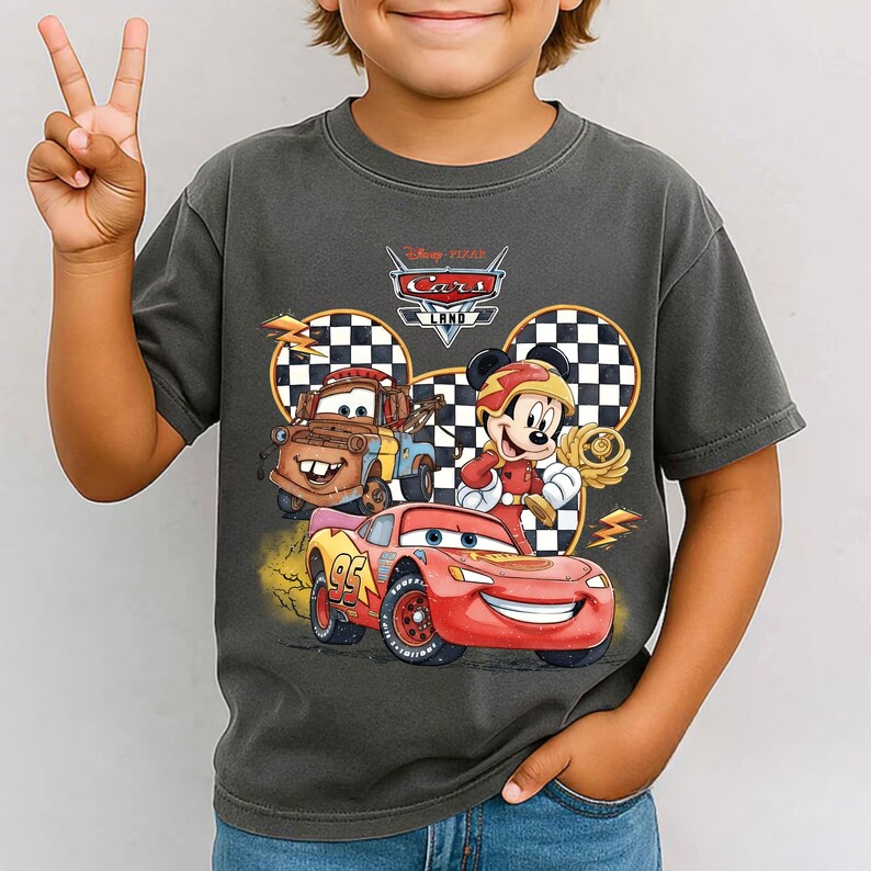 May include: A charcoal gray short-sleeve t-shirt featuring a graphic print of Disney Pixar Cars Land. The design includes Lightning McQueen, Mater, and Mickey Mouse in a racing outfit, set against a checkered background with yellow lightning bolts.