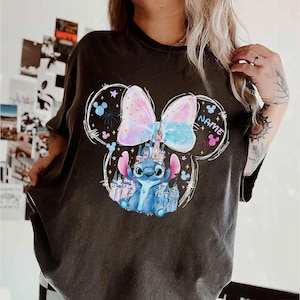 Pu&ograve; includere: T-shirt grigio scuro con un personaggio dei cartoni animati e un castello. Il fiocco &egrave; rosa pastello e blu. Il personaggio &egrave; blu con grandi occhi. La parola "NAME" &egrave; stampata sulla maglietta.