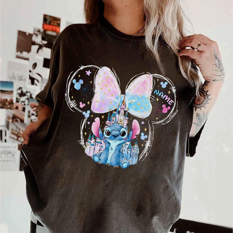 Pu&ograve; includere: T-shirt grigio scuro con un personaggio dei cartoni animati e un castello. Il fiocco &egrave; rosa pastello e blu. Il personaggio &egrave; blu con grandi occhi. La parola "NAME" &egrave; stampata sulla maglietta.