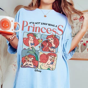Puede incluir: Una camiseta azul claro con un estampado gr&aacute;fico de Ariel de La Sirenita en cuatro paneles distintos. El texto superior dice IT'S NOT EASY BEING A PRINCESS en letras rojas y negras, con el logotipo de Disney impreso en la parte inferior.