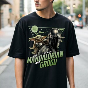 May include: A black short-sleeve t-shirt featuring a graphic print of The Mandalorian and Grogu. The design includes a green planet, a triangular frame, and the text "STAR WARS THE MANDALORIAN AND GROGU" in bold green lettering on the front.