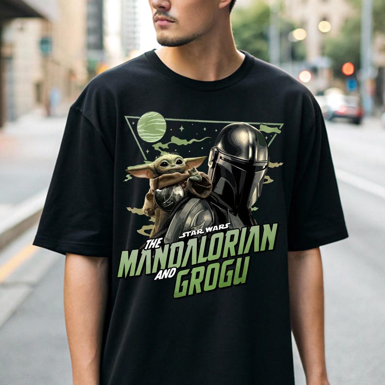 May include: A black short-sleeve t-shirt featuring a graphic print of The Mandalorian and Grogu. The design includes a green planet, a triangular frame, and the text "STAR WARS THE MANDALORIAN AND GROGU" in bold green lettering on the front.