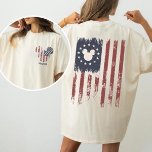 May include: Cream-colored t-shirt featuring a distressed American flag design on the back and a Mickey Mouse-themed flag on the front. The front design includes the word "NAME". The shirt is a casual, oversized fit.