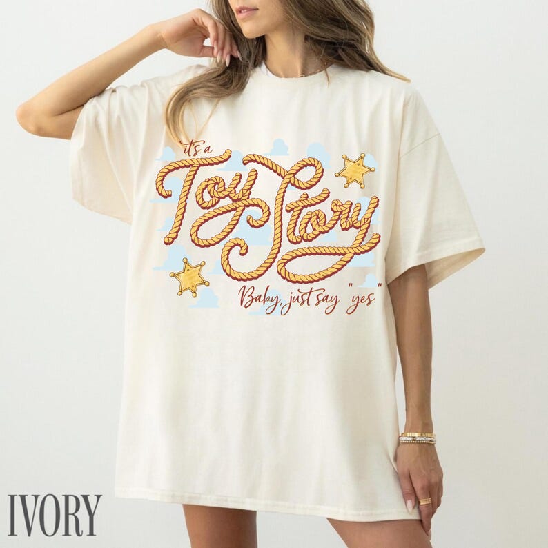 Peut inclure: Un t-shirt &agrave; manches courtes ivoire avec une impression graphique du texte "It's a Toy Story" &eacute;crit dans une police ressemblant &agrave; une corde, accompagn&eacute; de deux &eacute;toiles de sh&eacute;rif jaunes et de nuages bleu clair, avec le texte "Baby, just say yes".