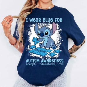 K&ouml;nnte beinhalten: Marineblaues T-Shirt mit Stitch, der ein Puzzleteil h&auml;lt. Der Text auf dem Shirt lautet: "I wear blue for Autism Awareness accept, understand, love."