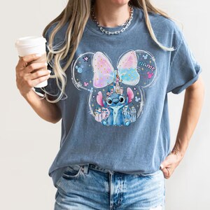Pu&ograve; includere: T-shirt blu con grafica di Stitch di Lilo & Stitch, un castello e un fiocco dai colori pastello. Il design include la parola "NAME" e le orecchie di Topolino.