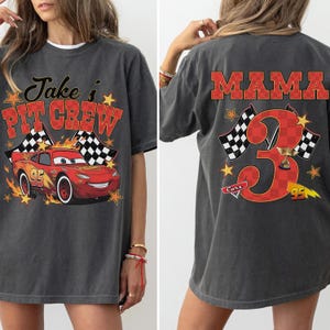 Puede incluir: Una camiseta gris carb&oacute;n con un gr&aacute;fico frontal de un coche de carreras rojo con el texto Jake's PIT CREW y un gr&aacute;fico trasero con un n&uacute;mero 3 grande y el texto MAMA, ambos dise&ntilde;os incluyen banderas a cuadros, estrellas y motivos de carreras.