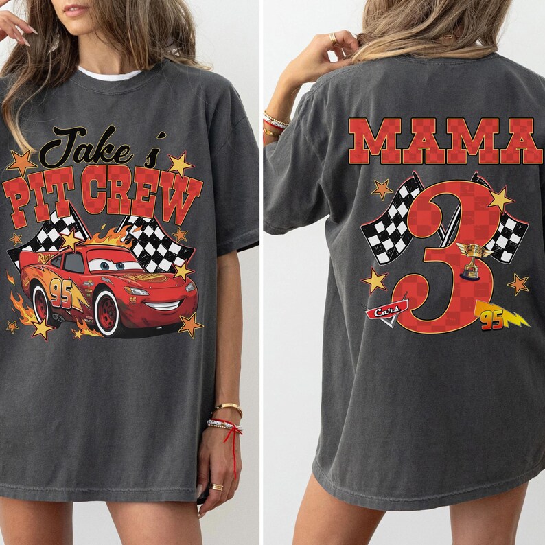 Puede incluir: Una camiseta gris carb&oacute;n con un gr&aacute;fico frontal de un coche de carreras rojo con el texto Jake's PIT CREW y un gr&aacute;fico trasero con un n&uacute;mero 3 grande y el texto MAMA, ambos dise&ntilde;os incluyen banderas a cuadros, estrellas y motivos de carreras.