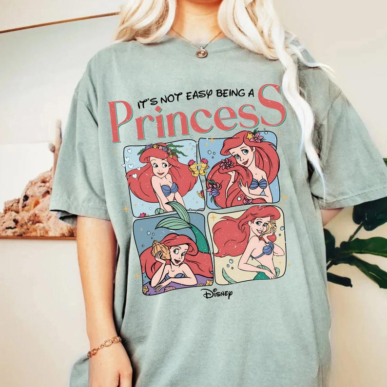 Puede incluir: Una camiseta de manga corta en color verde salvia con un estampado gr&aacute;fico que incluye el texto 'IT'S NOT EASY BEING A Princess' y cuatro paneles distintos que muestran al personaje Ariel de La Sirenita en varias poses con pelo rojo.