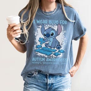 K&ouml;nnte beinhalten: Blaues T-Shirt mit dem Text "I wear blue for Autism Awareness" und einer Cartoonfigur, die ein Puzzleteil h&auml;lt. Das Shirt hat auch die Worte "accept, understand, love".