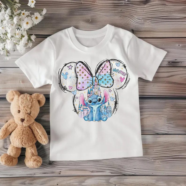 Pu&ograve; includere: T-shirt bianca con un cartone animato di un personaggio alieno blu, un castello e un fiocco. Il fiocco &egrave; rosa e blu a pois. La parola "NAME" &egrave; scritta sul disegno.