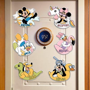 Peut inclure: Six autocollants muraux décoratifs représentant des personnages Disney sur des bouées, disposés sur une porte avec un panneau circulaire central indiquant 9152. Les personnages incluent Mickey nommé KEVIN, Minnie nommé SOPHIA, Donald nommé JACK, Daisy nommé SENA, Pluto nommé ZOO et Dingo nommé KEVIN.