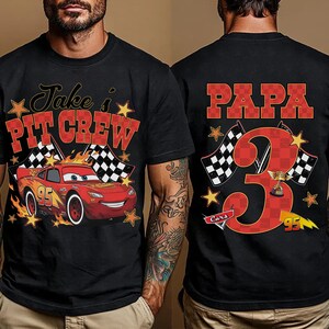 Puede incluir: Una camiseta negra con un gr&aacute;fico de un coche de carreras rojo con el n&uacute;mero 95, banderas a cuadros, estrellas amarillas y el texto Jake's PIT CREW en el frente. La parte trasera muestra el texto PAPA, un n&uacute;mero 3 grande y un trofeo.