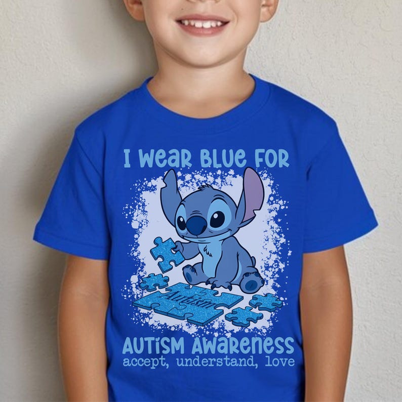 K&ouml;nnte beinhalten: Ein k&ouml;nigsblaues T-Shirt mit einer Cartoonfigur, die ein Puzzleteil h&auml;lt. Das Shirt tr&auml;gt den Text "I wear blue for Autism Awareness" mit den Worten "accept, understand, love" unter der Grafik. Das Shirt wird von einem Kind getragen.