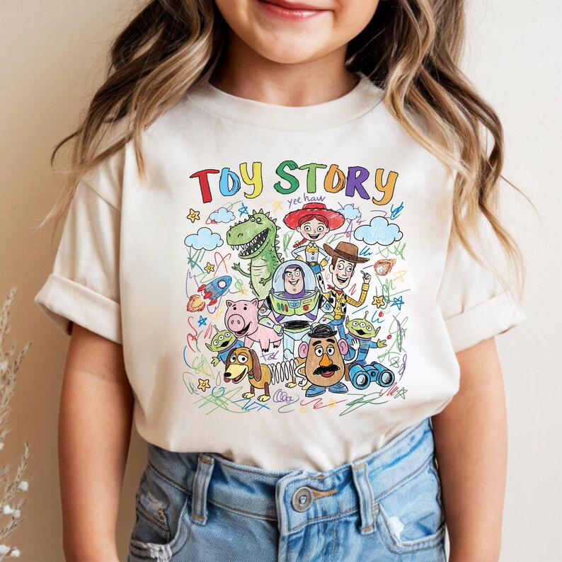 May include: A cream-coloured t-shirt with a vibrant graphic of characters from the animated film "Toy Story." The design includes the words "Toy Story" in a playful font, along with images of Woody, Buzz Lightyear, and other characters.