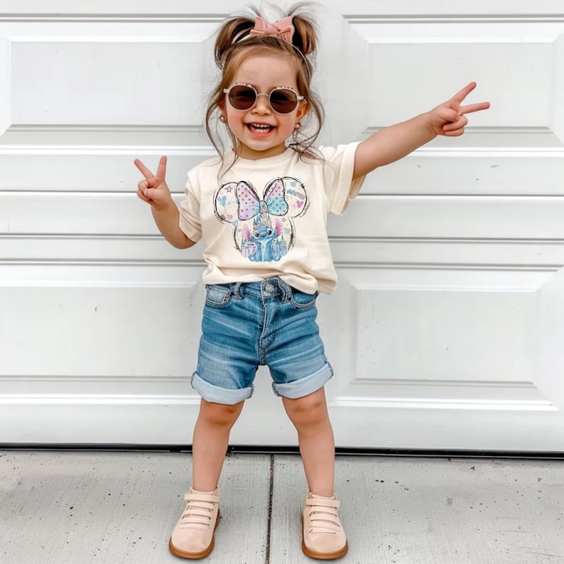 Pu&ograve; includere: Una bambina indossa una maglietta color crema con una grafica di Minnie Mouse, pantaloncini di jeans e scarpe color cuoio. La grafica di Minnie Mouse presenta un fiocco e il personaggio Stitch. La bambina indossa occhiali da sole e fa il segno della pace.