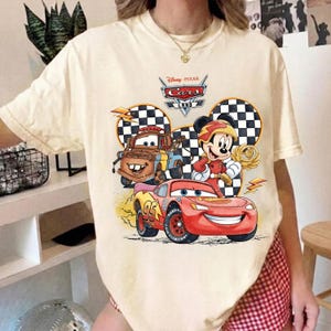 May include: A cream-colored t-shirt featuring a graphic print of Disney Pixar Cars characters Lightning McQueen and Mater alongside Mickey Mouse. The design includes a checkered pattern, lightning bolts, and the text Disney Pixar Cars Land.