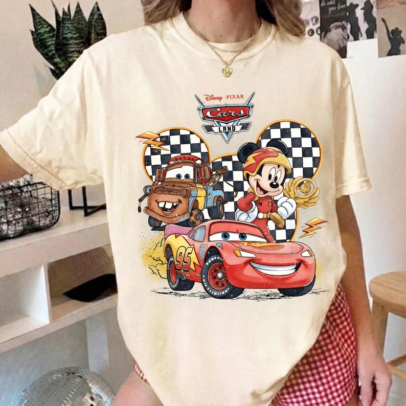 May include: A cream-colored t-shirt featuring a graphic print of Disney Pixar Cars characters Lightning McQueen and Mater alongside Mickey Mouse. The design includes a checkered pattern, lightning bolts, and the text Disney Pixar Cars Land.