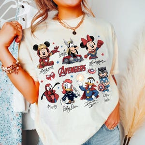 May include: A cream-coloured t-shirt with a vibrant graphic of Disney characters reimagined as Avengers superheroes. The design includes Mickey Mouse, Minnie Mouse, Goofy, Pluto, Daisy Duck, Donald Duck, and Chip and Dale. The word "Avengers" is displayed.