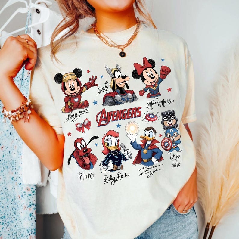 May include: A cream-coloured t-shirt with a vibrant graphic of Disney characters reimagined as Avengers superheroes. The design includes Mickey Mouse, Minnie Mouse, Goofy, Pluto, Daisy Duck, Donald Duck, and Chip and Dale. The word "Avengers" is displayed.