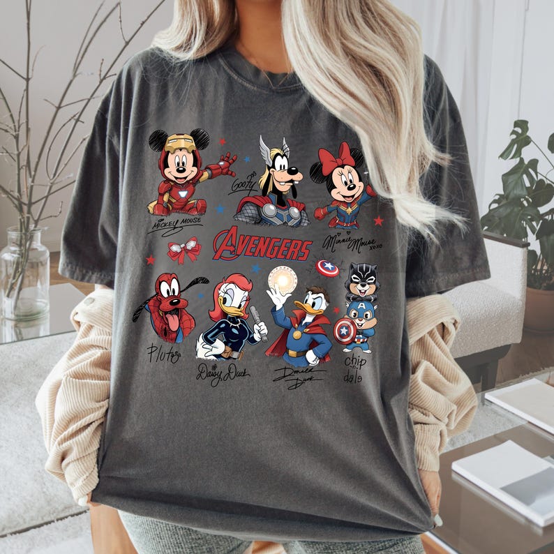 May include: Dark grey t-shirt with a vibrant graphic of Disney characters styled as Avengers superheroes. The design includes Mickey Mouse, Minnie Mouse, Goofy, Pluto, Daisy Duck, Donald Duck, and Chip and Dale. The word "Avengers" is also displayed.