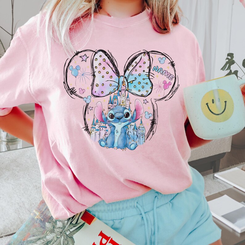 Pu&ograve; includere: T-shirt rosa con un'immagine a cartoni animati di Stitch di Lilo & Stitch all'interno di un contorno della testa di Topolino. Il design include un castello, un fiocco e la parola "NAME". La persona tiene in mano una tazza azzurra con una faccina gialla.
